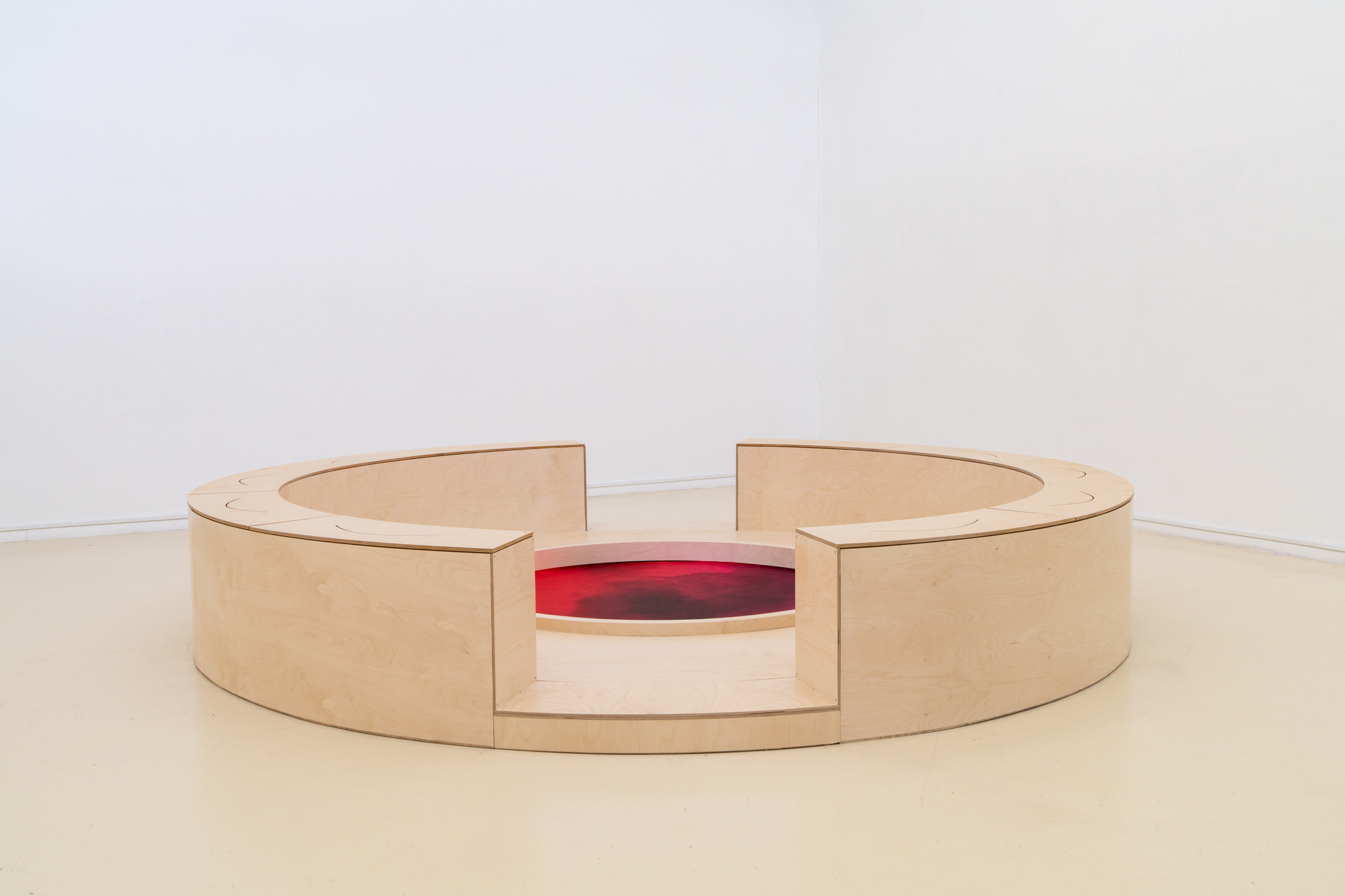 Single-Plural Body seen from its entrance: an empty circular plywood seating ring with a dark red screen set into the floor