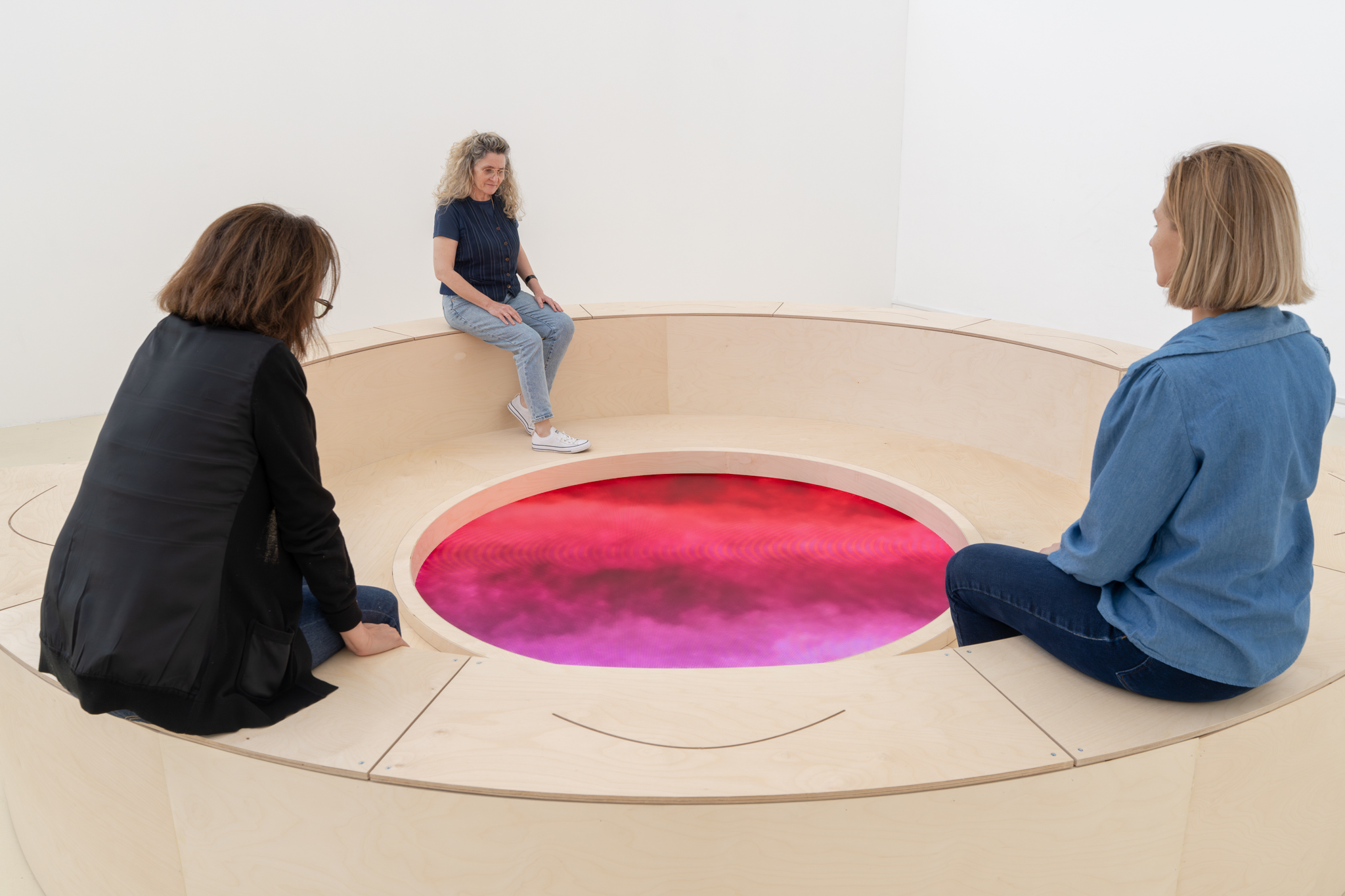 Three visitors seated around the installation, looking down at a pink, cloud-like image on the floor screen